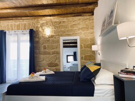 Un dormitorio con pared de piedra, una cama con cubrecama oscuro, una mesita de noche con lámpara y una ventana con cortinas.