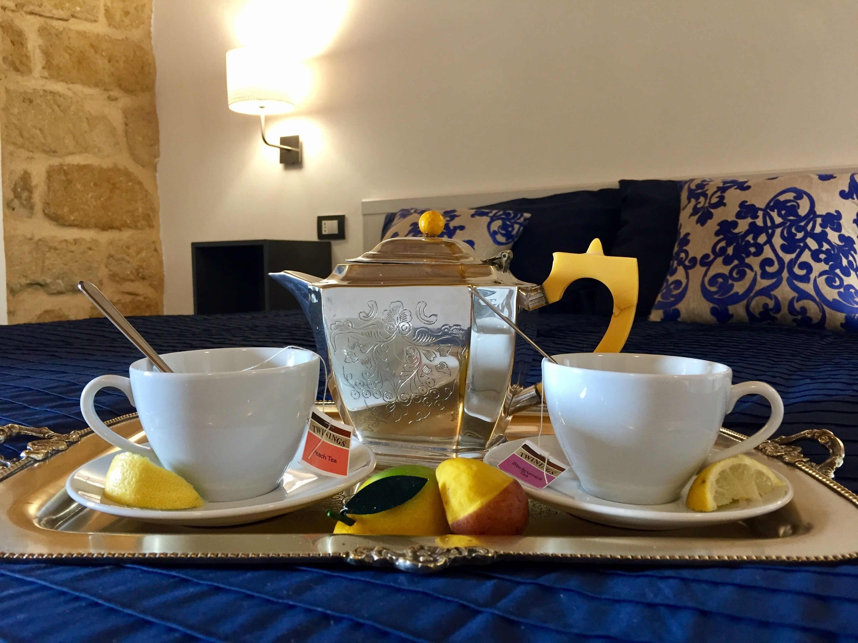 A breakfast tray with two white cups, a teapot, and fruit on a bed with a blue bedspread.