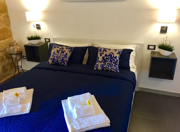 Una cama bien tendida con ropa de cama azul y almohadas decorativas, dos mesitas de noche con lámparas y plantas, y una pared de piedra decorativa.