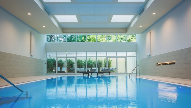 An indoor swimming pool with clear glass walls, a tiled floor, and a few lounge chairs. - Eilenriedestift Hotel (Hannover)