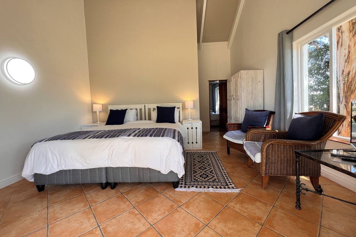 Een hotelkamer met een groot bed, twee nachtkastjes met lampen, een rieten stoel, een klein tafeltje en uitzicht op het omliggende landschap door een raam.
