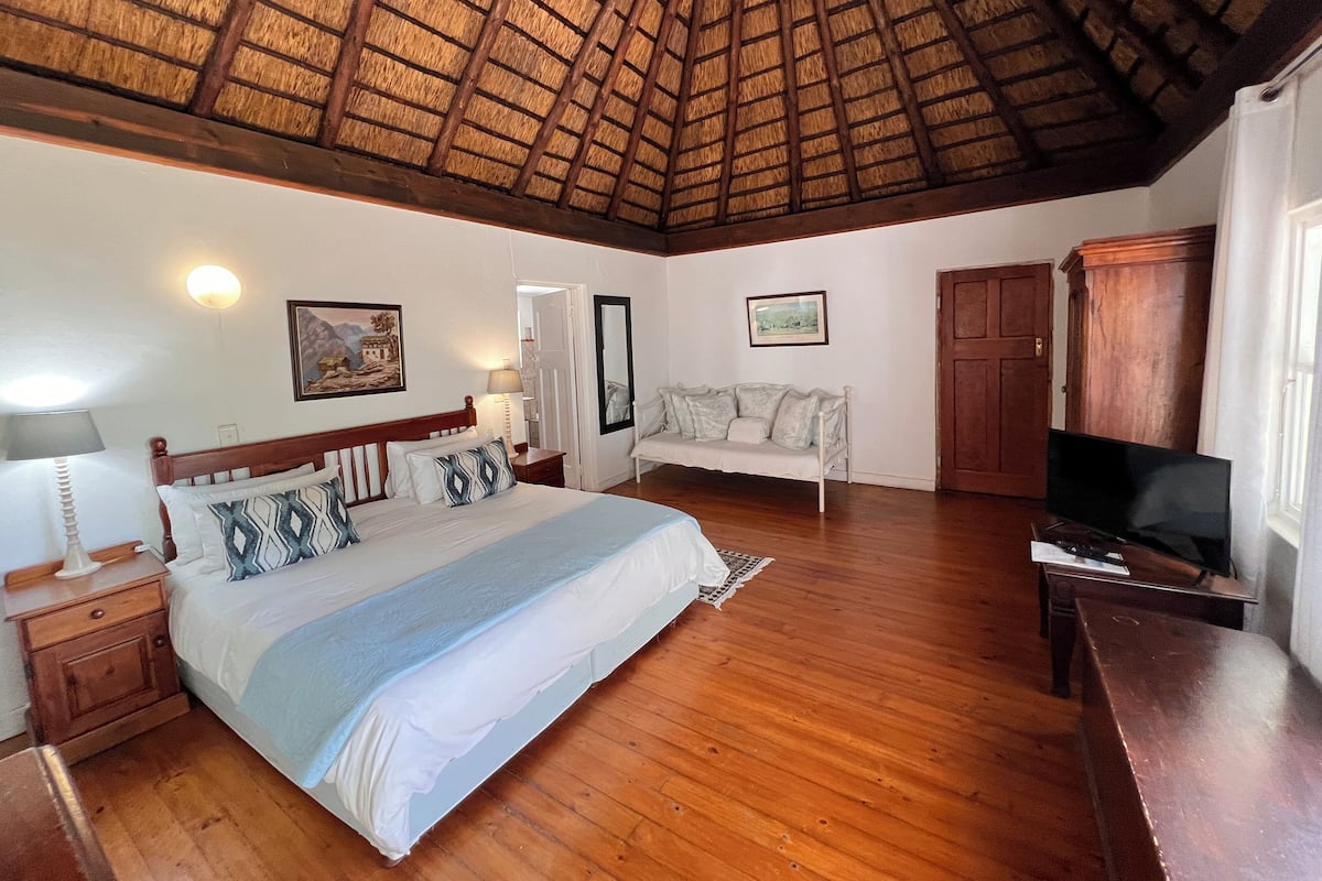 Een ruime slaapkamer met een houten plafond, een bed met gestreepte kussens, een bank, een televisie en een kledingkast.