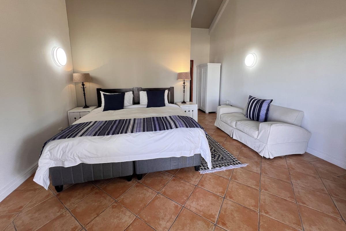 Een slaapkamer met een groot bed, een nachtkastje met een lamp, een witte kledingkast en een bank met een decoratief kussen.