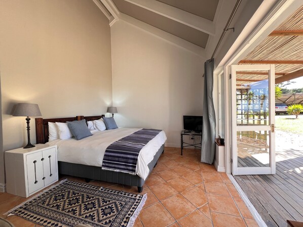 A bedroom with a bed, bedside lamps, a TV, and a view of the outside. - The Whaling Station Guesthouse (Plettenberg Bay)