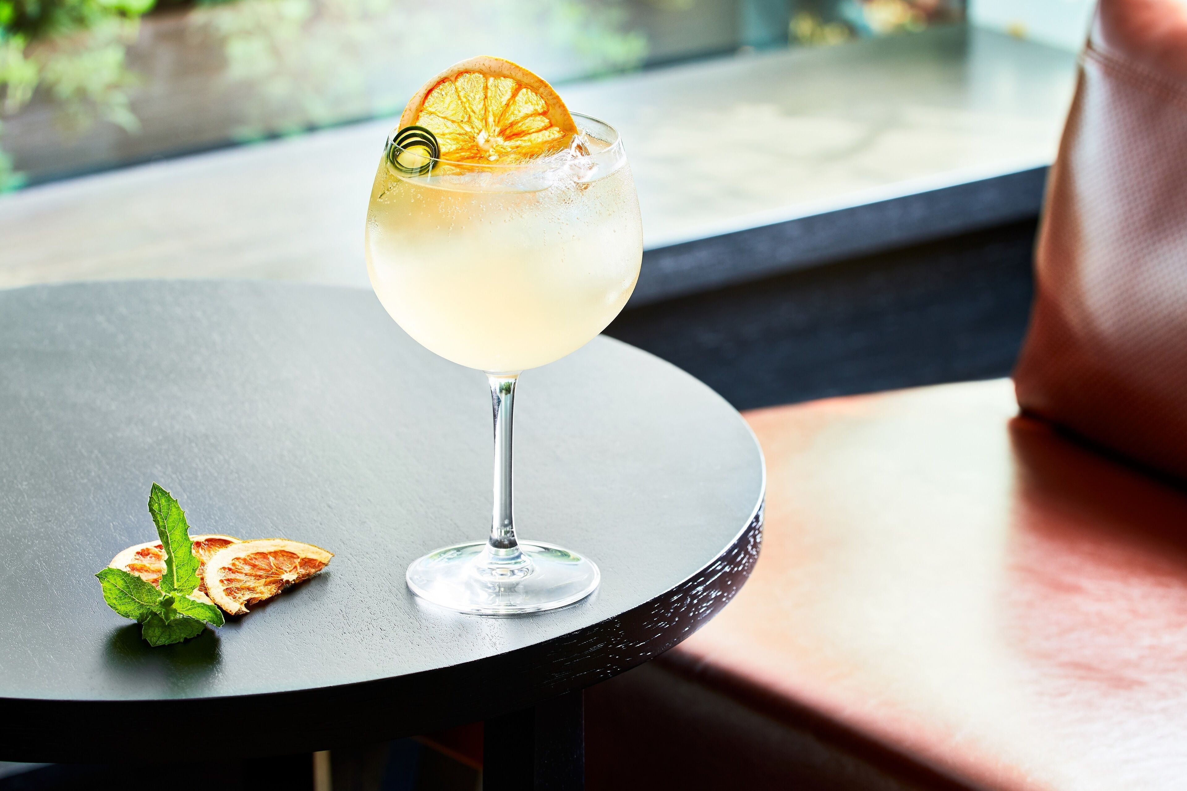 Un verre à cocktail orné d’une tranche d’orange sur le bord et de quelques feuilles de menthe, posé sur une table ronde.