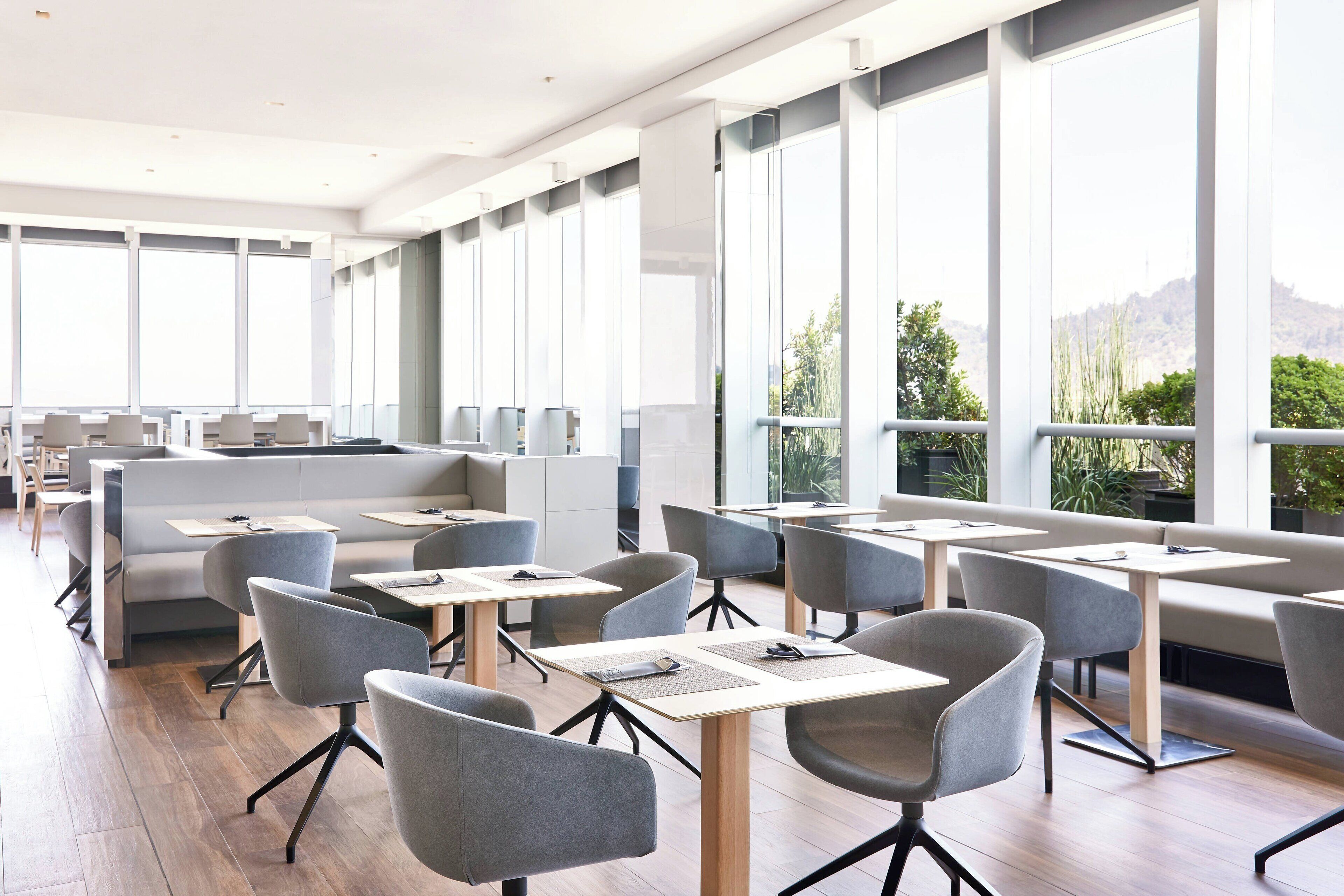 A modern dining area with high tables and chairs, large windows, and a view of greenery and hills.