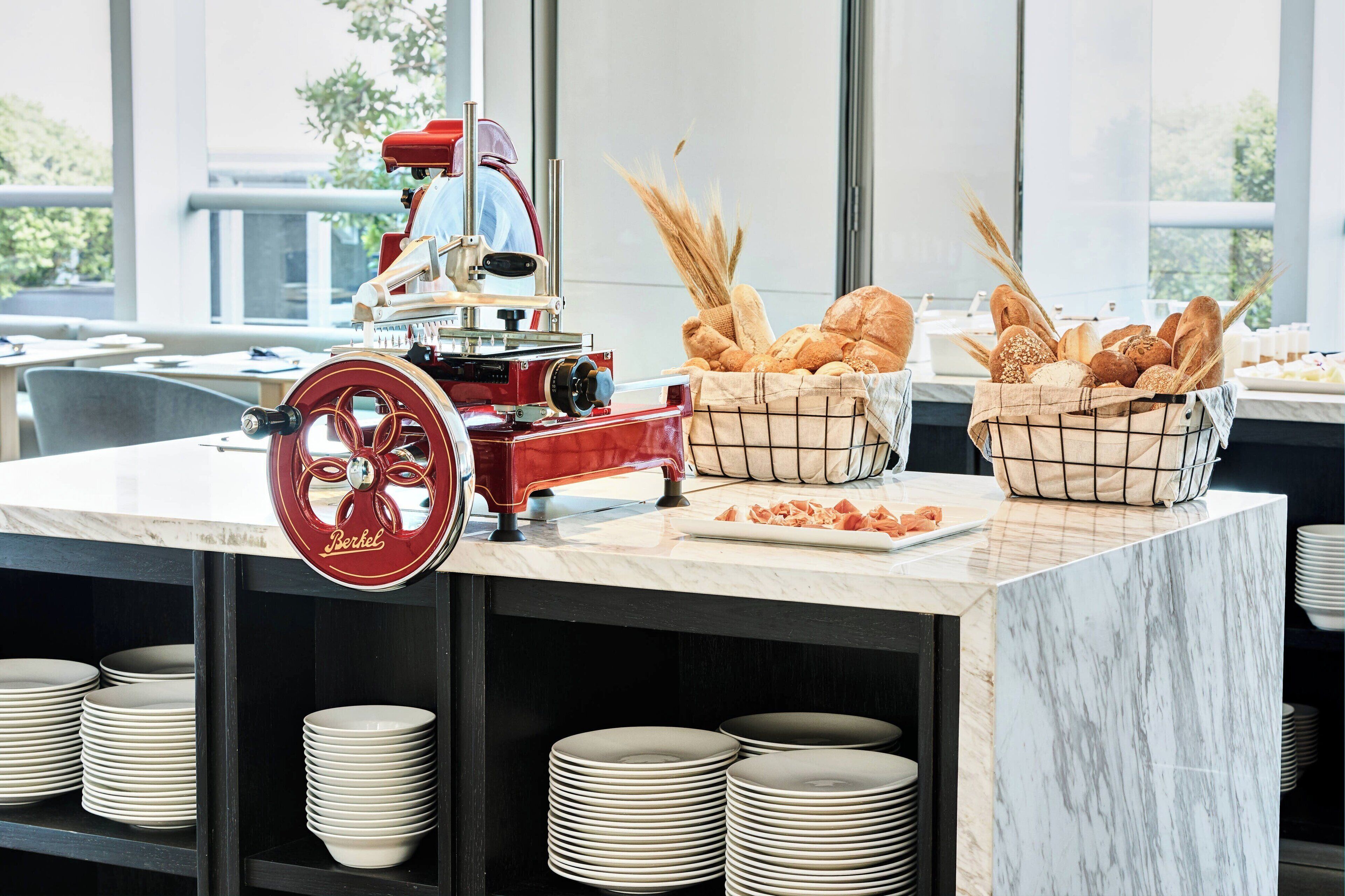 Una mesa de buffet con un procesador de alimentos rojo, cestas de pan y platos apilados.