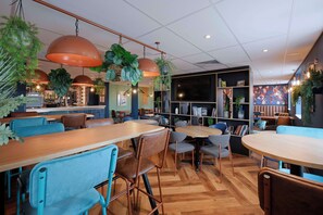 A modern cafe with wooden tables and chairs, hanging plants, and a bookshelf with books and decorative items. - Travelodge London Finchley (London)
