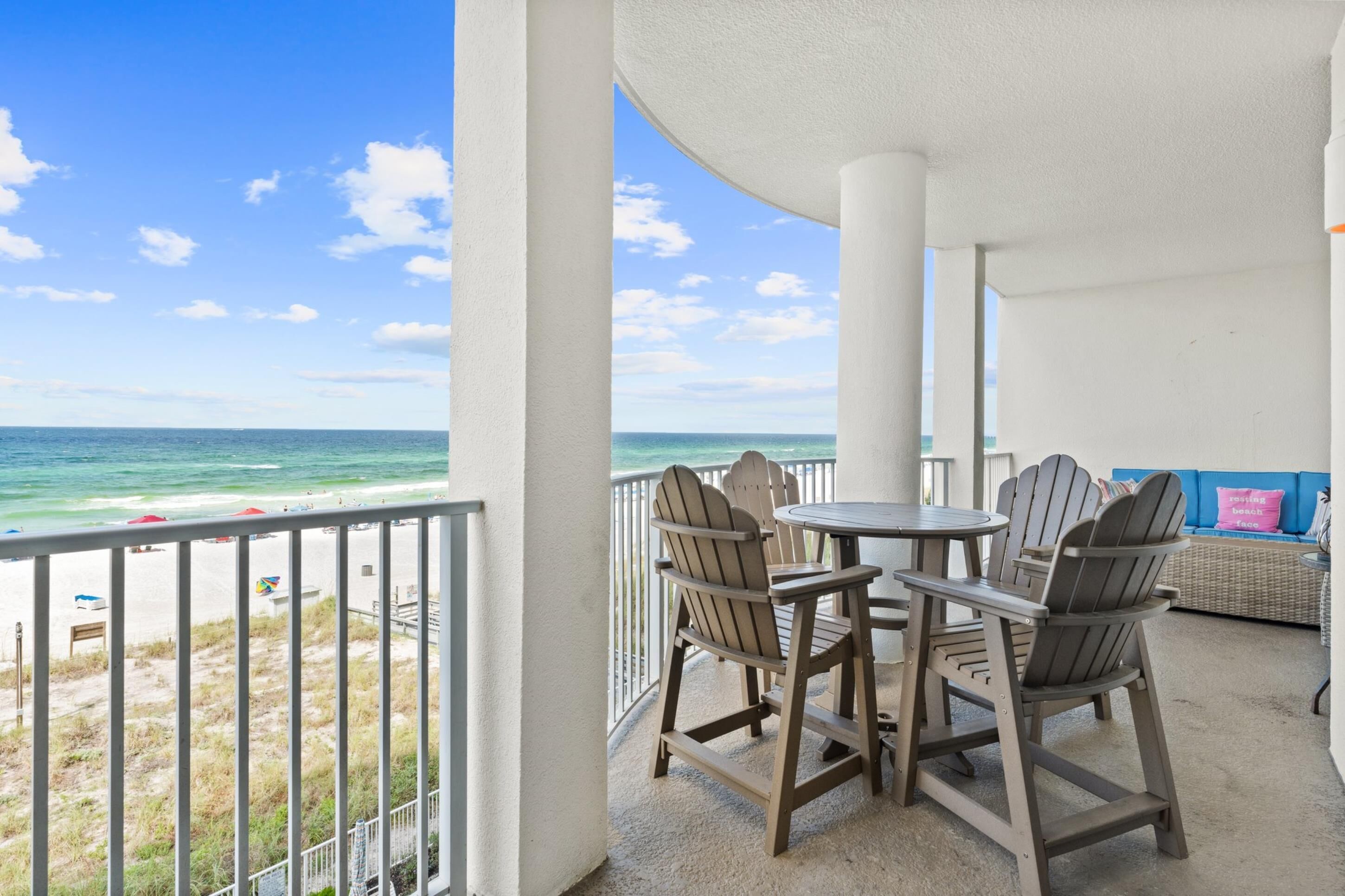 A balcony with a view of the beach and ocean, featuring Adirondack chairs and a round table.