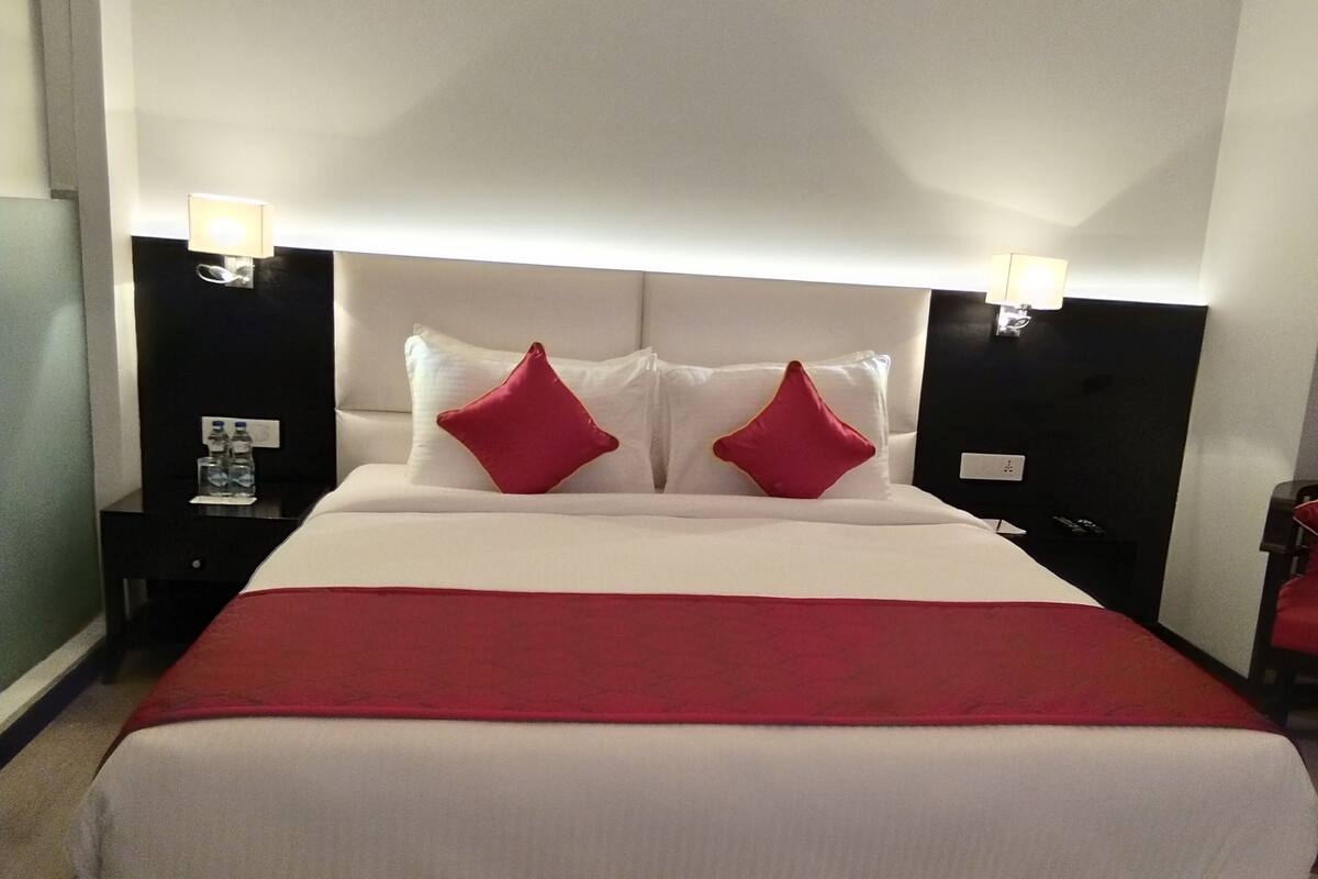 A hotel room with a large bed, two red pillows, a red throw blanket, and two bedside tables with lamps.