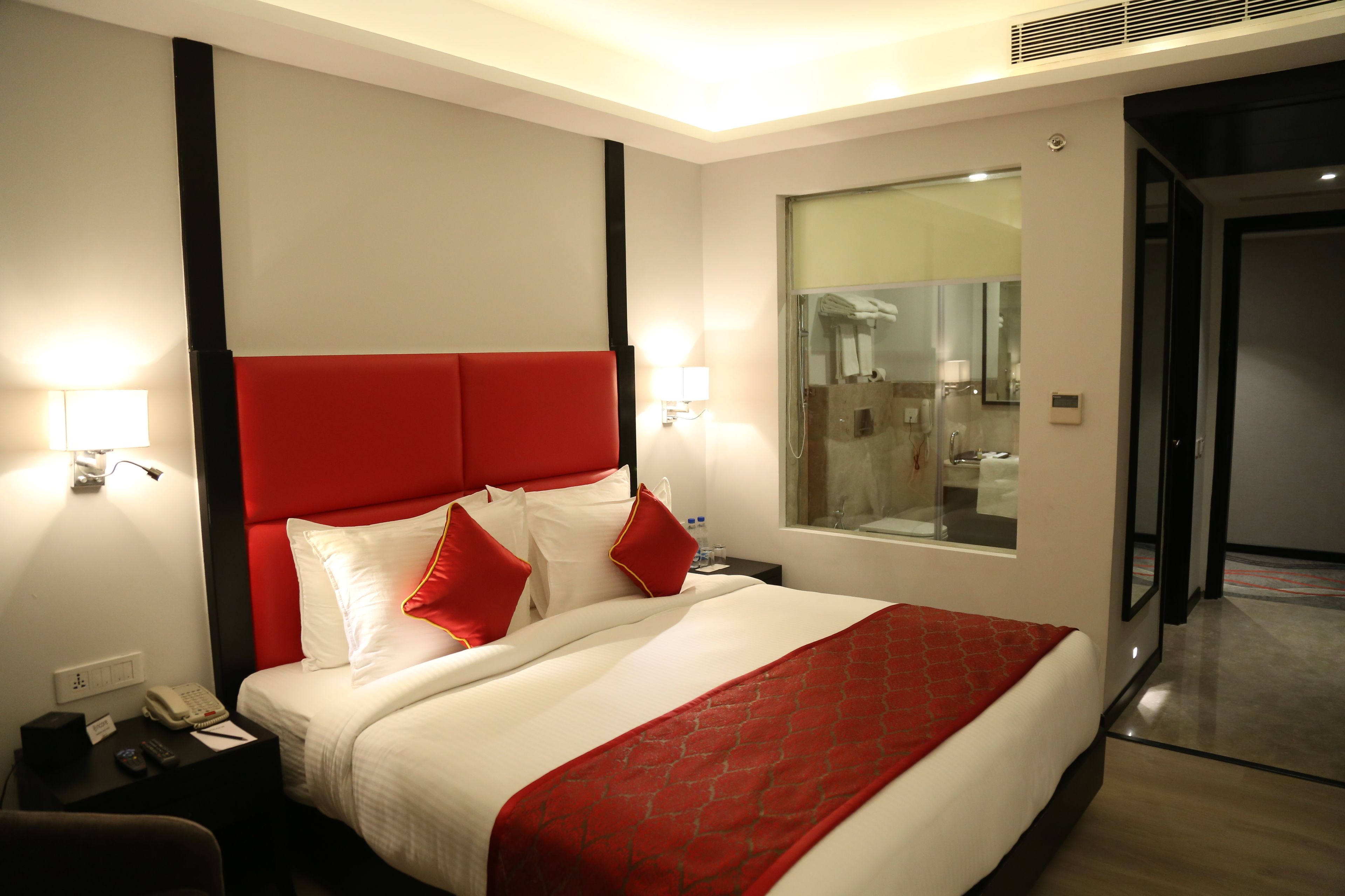 Une chambre d’hôtel avec un grand lit, une tête de lit rouge, du linge de lit blanc et un coussin décoratif rouge. On y trouve une table de chevet avec un téléphone et une lampe, ainsi qu’une fenêtre donnant sur la salle de bain.