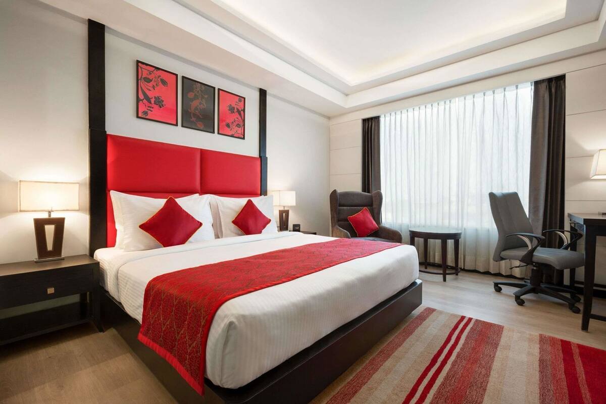 A hotel room with a large bed, a desk, two chairs, a window with curtains, and artwork on the wall.