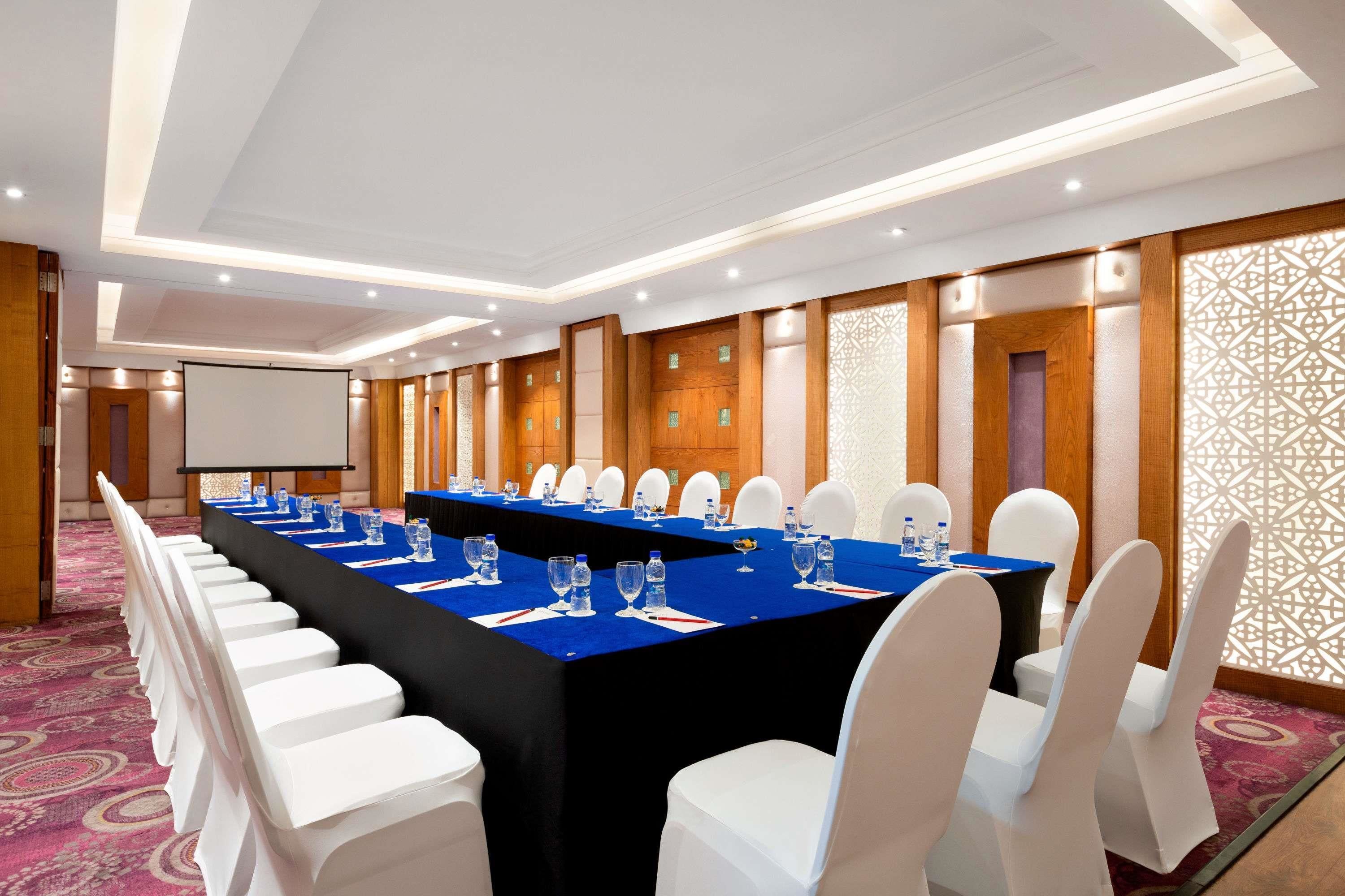Un salón de conferencias con una larga mesa, sillas blancas y una pantalla para proyector.