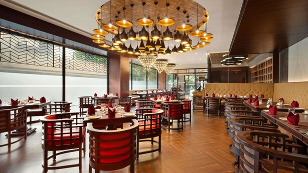 A restaurant with wooden tables and chairs, a large chandelier, and a view of the street through large windows. - Ramada Encore by Wyndham Jalandhar (Jalandhar)
