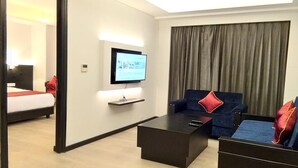 A modern hotel room with a flat-screen TV mounted on the wall, a dark-colored sofa with red cushions, a small coffee table, and a bed with a red accent pillow. - Ramada Encore by Wyndham Jalandhar (Jalandhar)