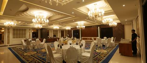 Un salón de banquetes con mesas redondas, sillas blancas, candelabros y una alfombra estampada.