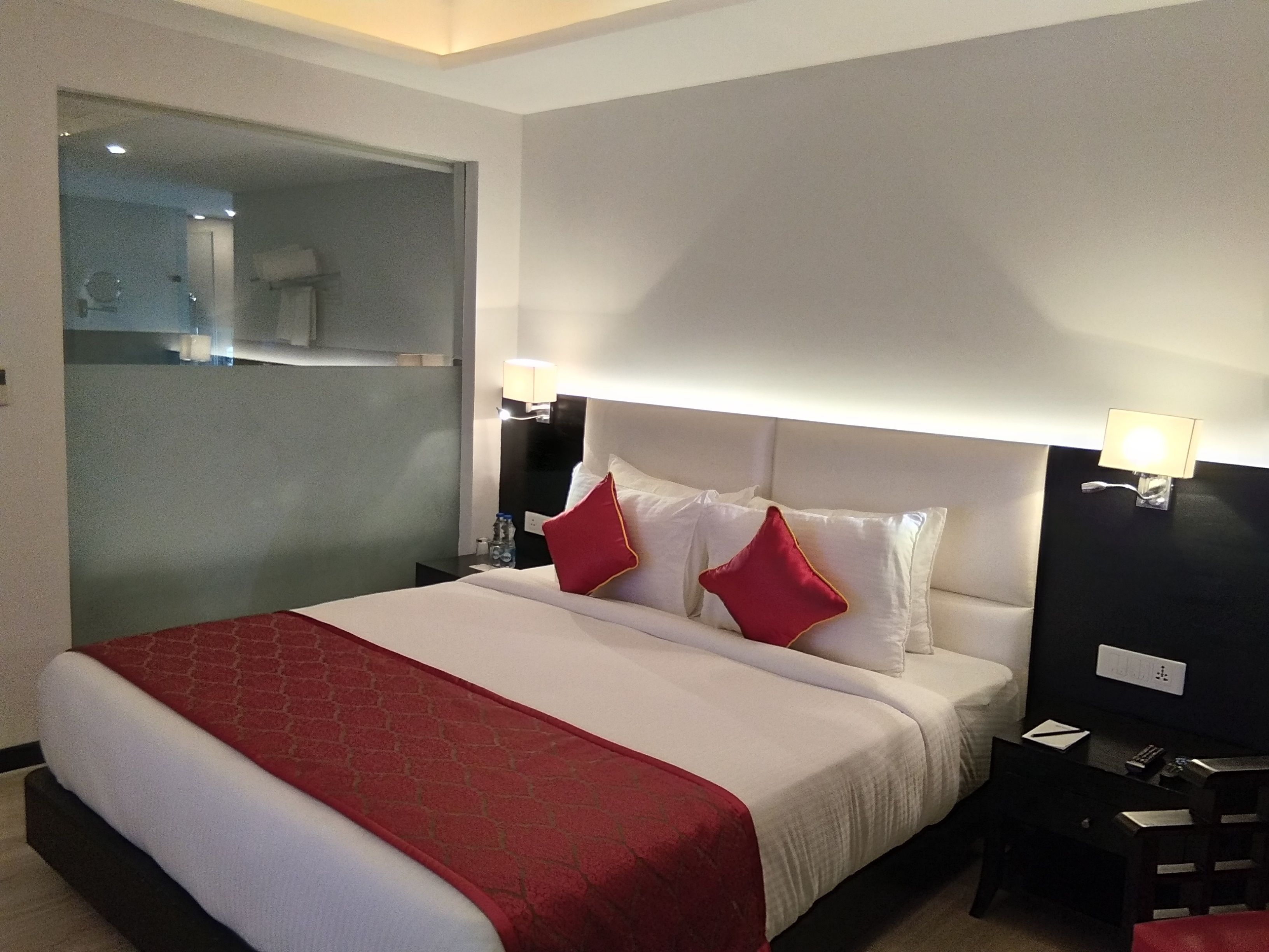 A hotel room with a large bed, two bedside lamps, a nightstand, and a glass-enclosed shower area.