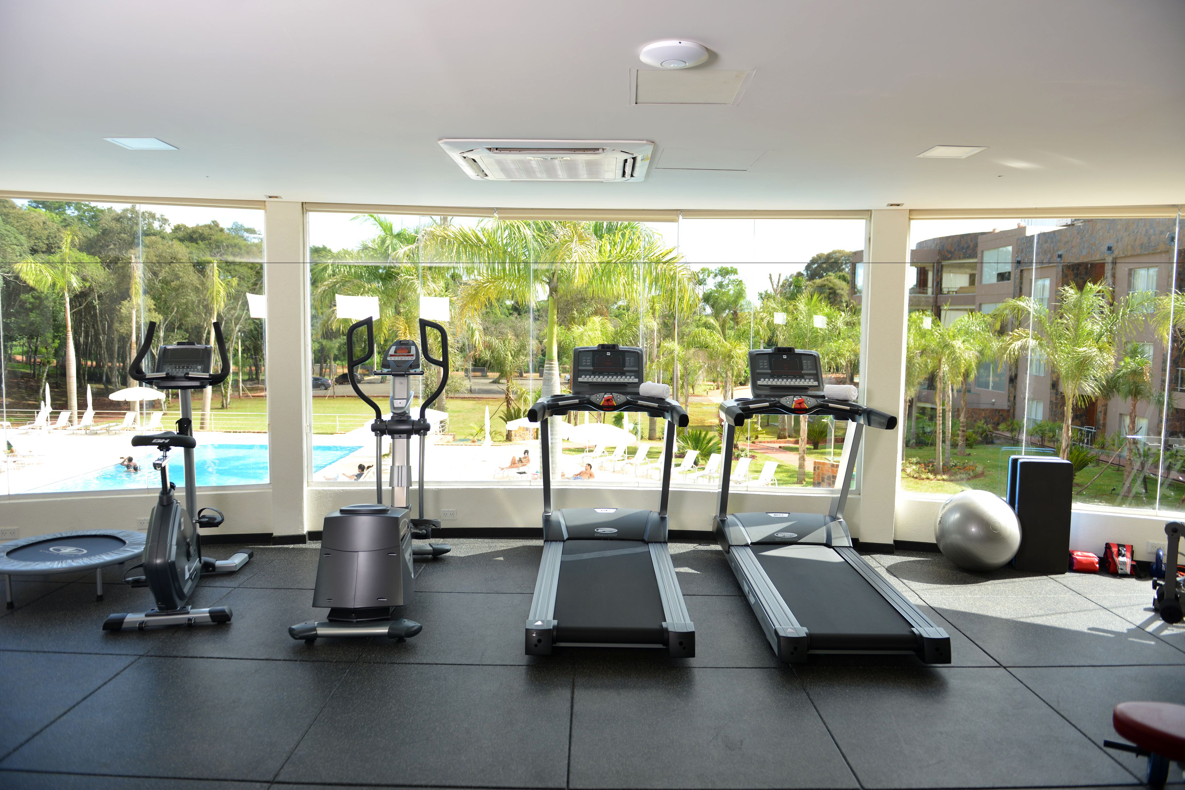 Un gimnasio con cintas de correr, elípticas y una bicicleta estática, con vistas a una piscina y abundante vegetación.