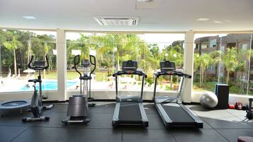 Un gimnasio con cintas de correr, elípticas y una bicicleta estática, con vistas a una piscina y abundante vegetación.