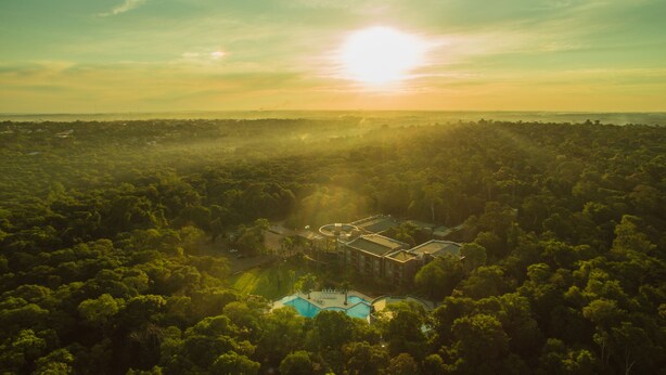 An aerial view of a resort with a swimming pool surrounded by dense forest, and a large building with a flat roof in the background.