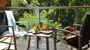 A balcony with two chairs, a table with breakfast items, and a view of dense greenery.