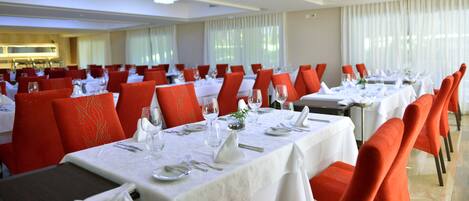 Un comedor con varias mesas dispuestas para el servicio, sillas tapizadas en rojo y un mantel blanco sobre cada una.