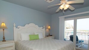 Une chambre à coucher avec un lit, deux tables de chevet avec des lampes, un ventilateur de plafond et une vue sur la plage, accessible par une porte-fenêtre coulissante.