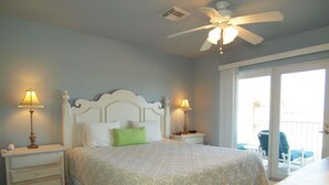 Une chambre à coucher comprenant un lit, deux tables de chevet avec des lampes, un ventilateur de plafond et une vue sur l’extérieur grâce à une porte-fenêtre coulissante.