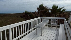Una terraza de madera con baranda, un banco y una silla con vista a una playa con palmeras y el océano.