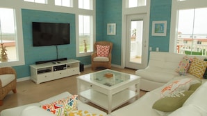A spacious living room with a large TV, white sofas, and a glass coffee table.
