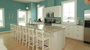 A kitchen with a large island, white bar stools, a stainless steel refrigerator, and a view through the window.