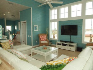 Ein geräumiges Wohnzimmer mit einem großen Fernseher, weißen Sofas und einem Deckenventilator.