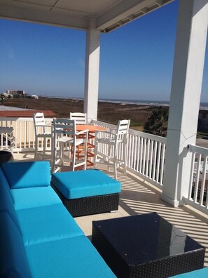 A covered outdoor seating area with blue cushioned sofas and a glass-top table, overlooking a beach.