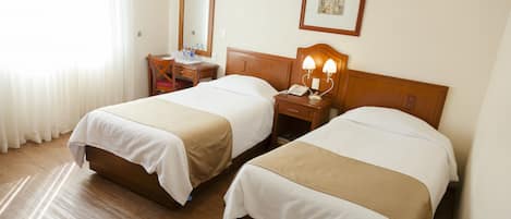 Quarto de hotel com duas camas, cabeceira de madeira, mesa de cabeceira com abajur, espelho e quadro na parede.