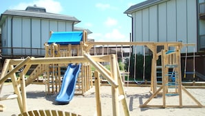 Une aire de jeux avec un toboggan et des balançoires, entourée d’une zone sablonneuse et de structures de jeu en bois.