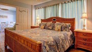 A bedroom with a wooden bed, patterned bedding, a ceiling fan, and a nightstand with a lamp. - Turtle Bay 104BH (Port Aransas)