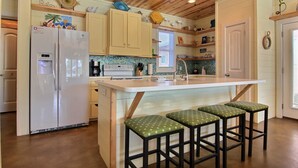 Una cocina con armarios de madera, un refrigerador blanco, una isla con taburetes y un ventanal con persianas.