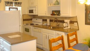 A compact kitchen with white cabinets, a stove, microwave, and a coffee maker. There are two wooden chairs and a small table with a plant. - Turtle s Pace SIV5 (Port Aransas)