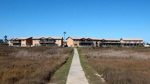 A long, straight pathway leading to a row of multi-story buildings with balconies, situated in a grassy area with a clear sky. - Turtle s Pace SIV5 (Port Aransas)
