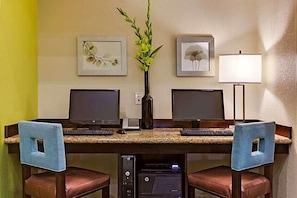 Ein Hotelzimmer mit Schreibtisch, zwei Monitoren, einer Lampe und einer Vase mit Blumen.