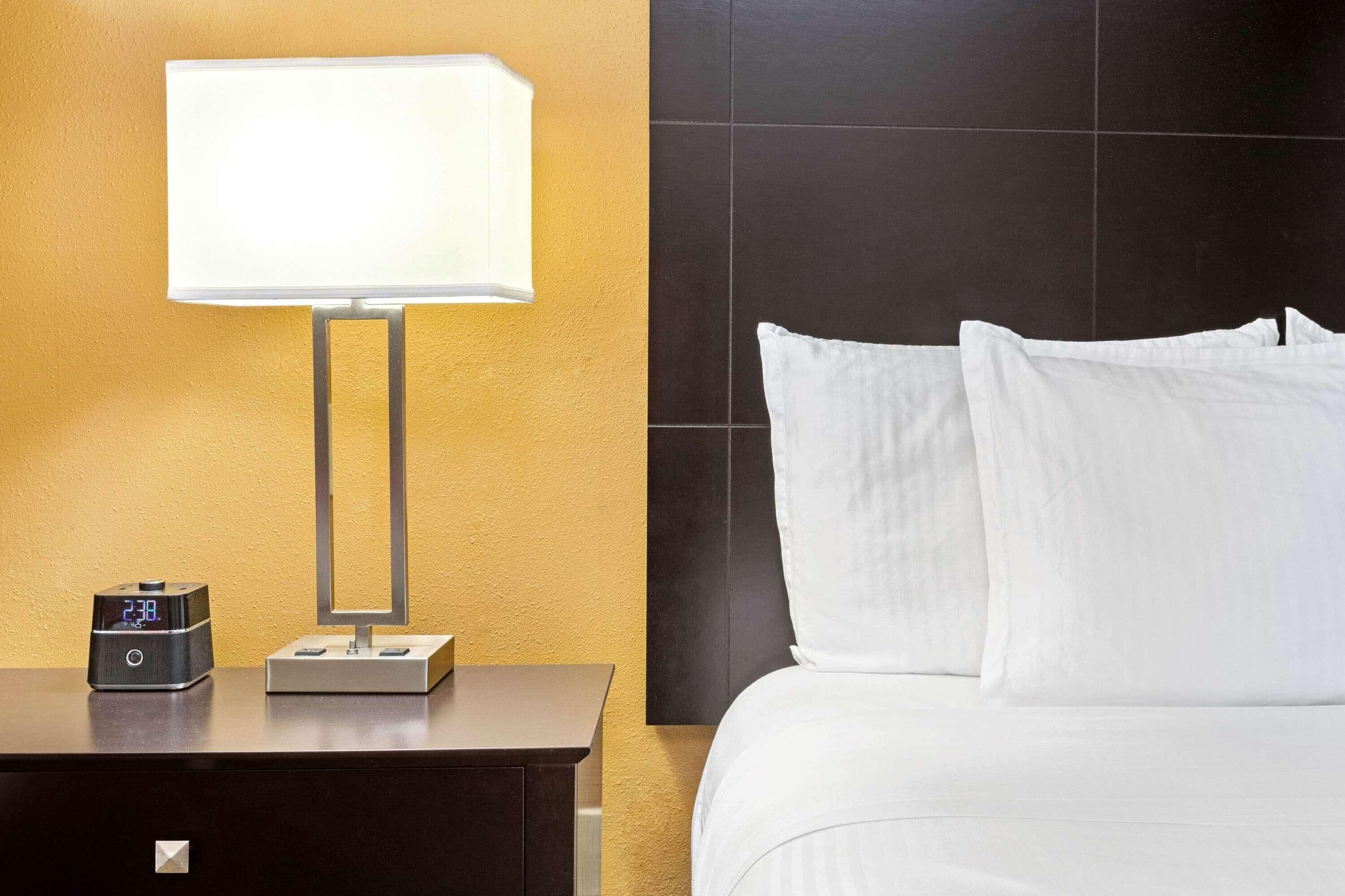 A hotel room with a bed, a nightstand, a digital clock, and a wall-mounted lamp.