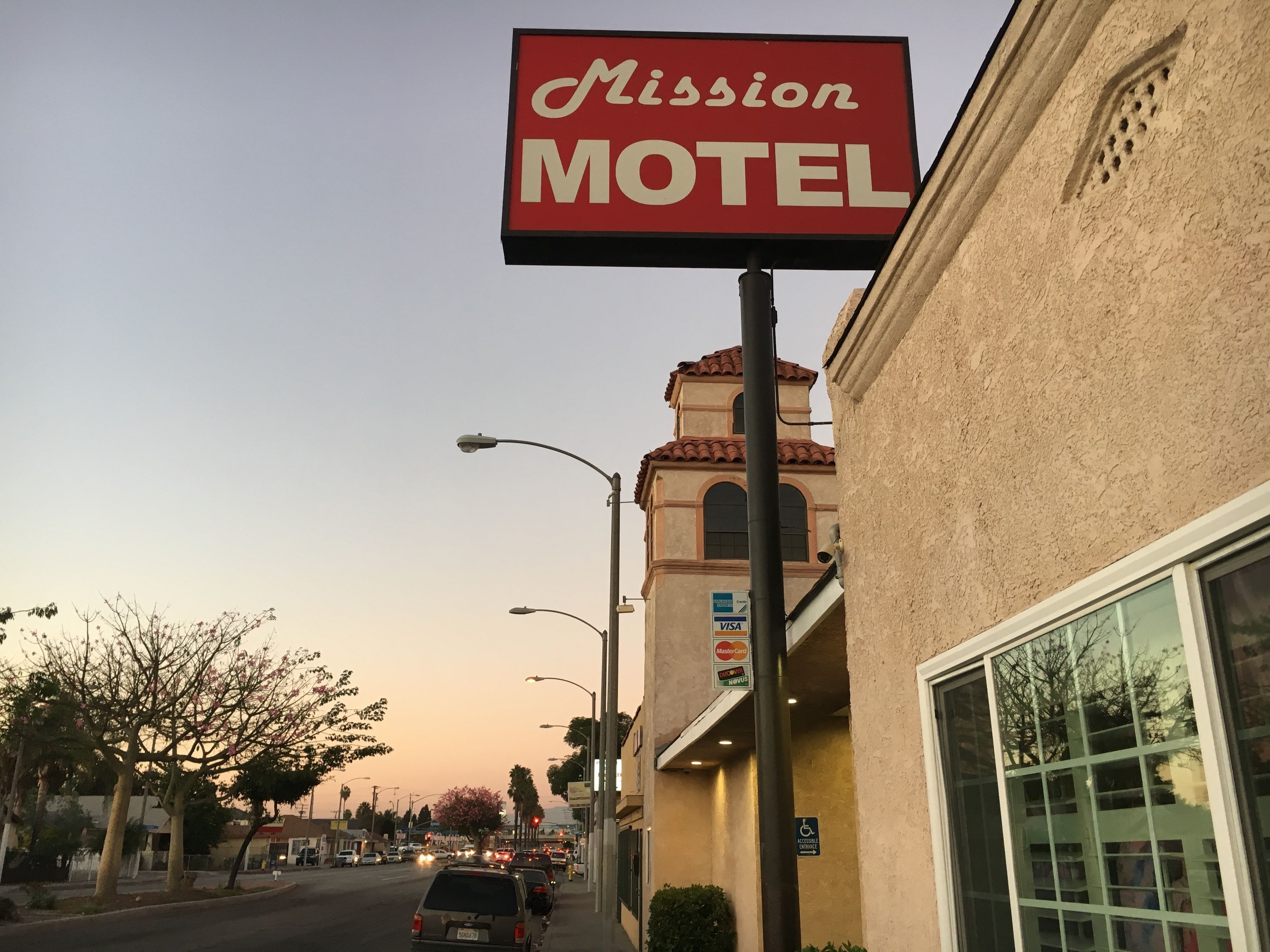 Katunäkymä, jossa näkyy kyltti, jossa lukee ”Mission Motel”, beige julkisivulla varustettu rakennus ja kirkas taivas.