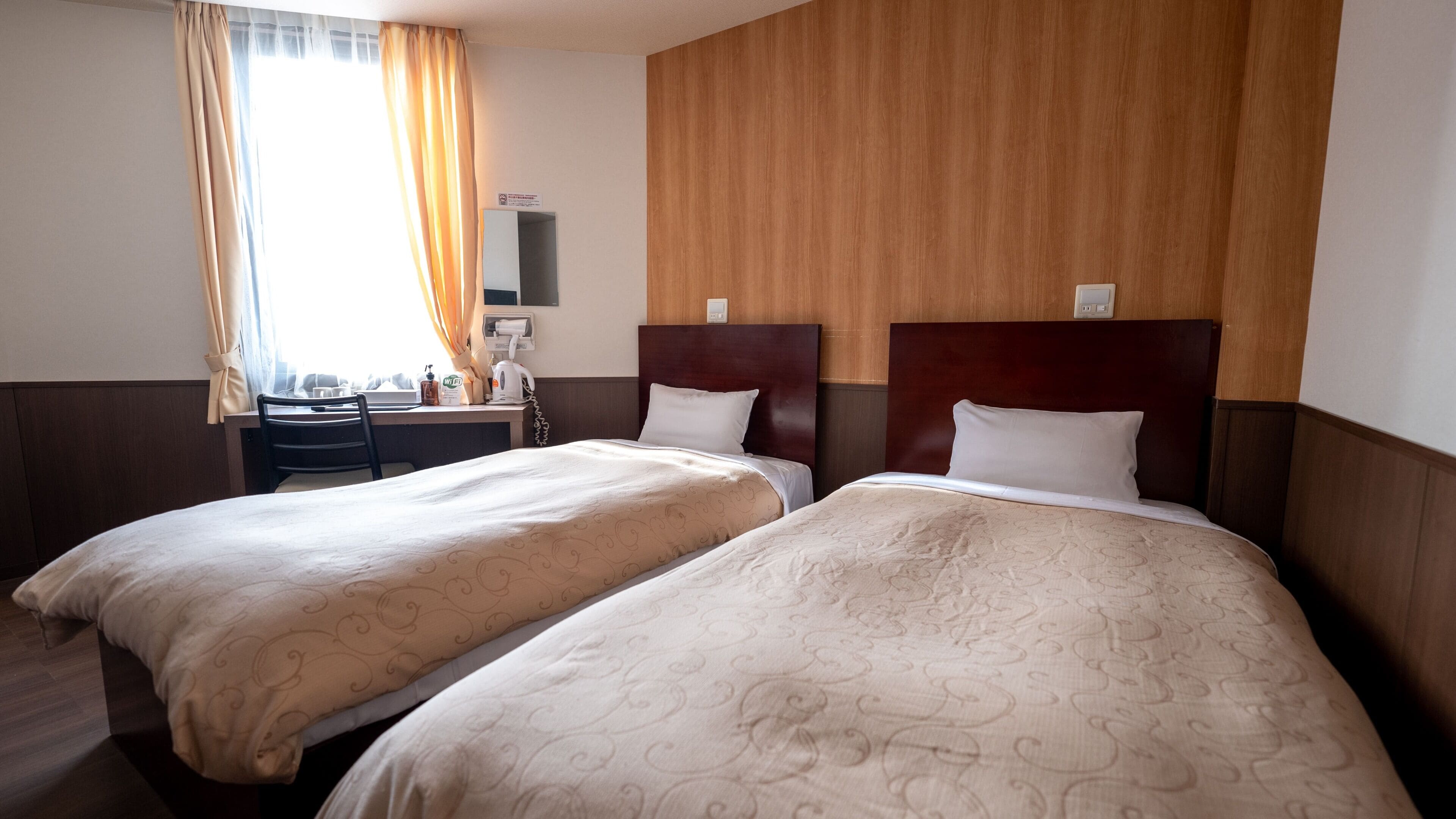 A hotel room with two beds, a desk, a chair, a window with curtains, and a wooden headboard.