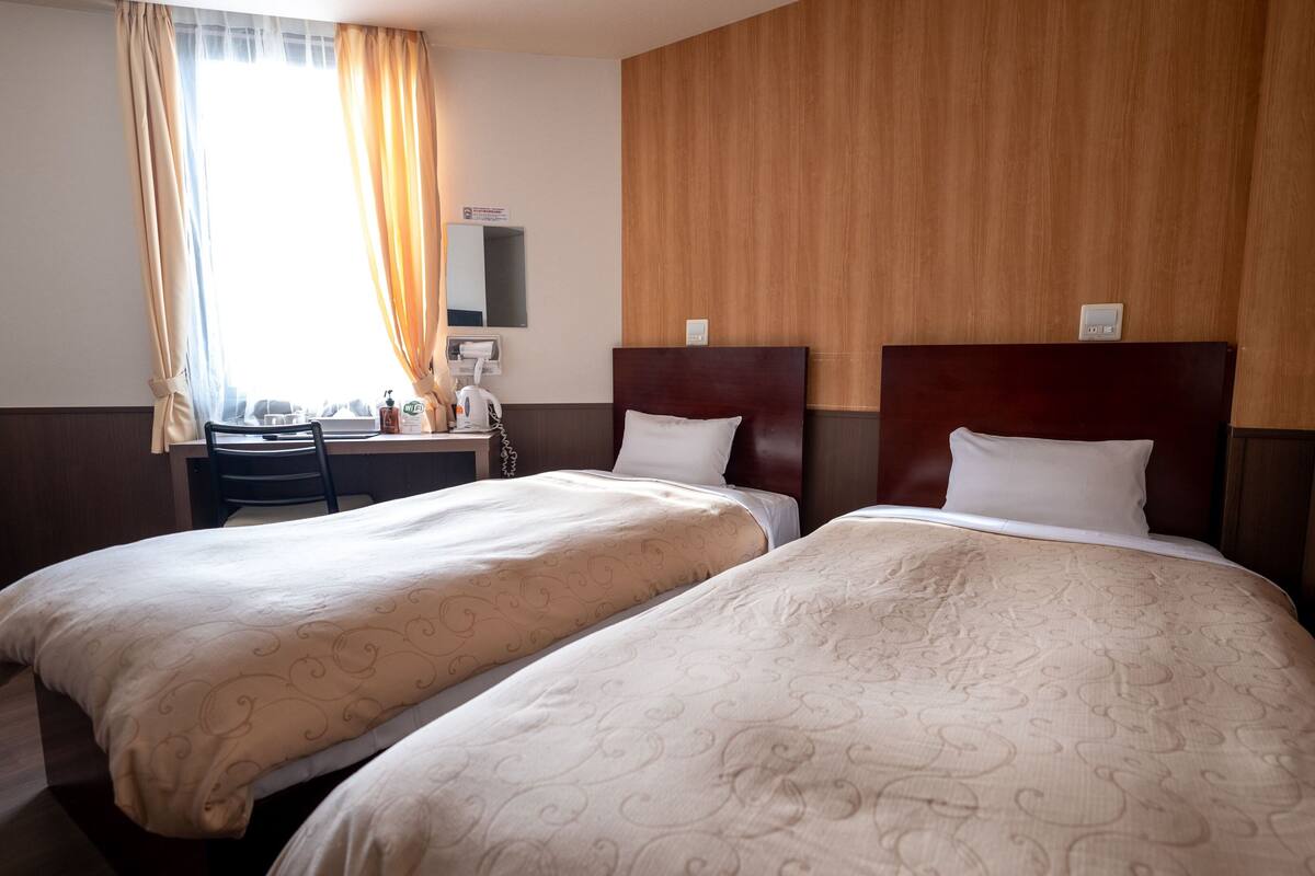 Habitación de hotel con dos camas, un escritorio, una silla, una ventana con cortinas y un cabecero de madera.