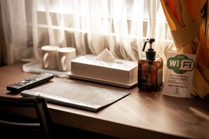 Escritorio de habitación de hotel con un control remoto, una caja de pañuelos, una botella de gel antibacterial y un cartel de Wi-Fi.
