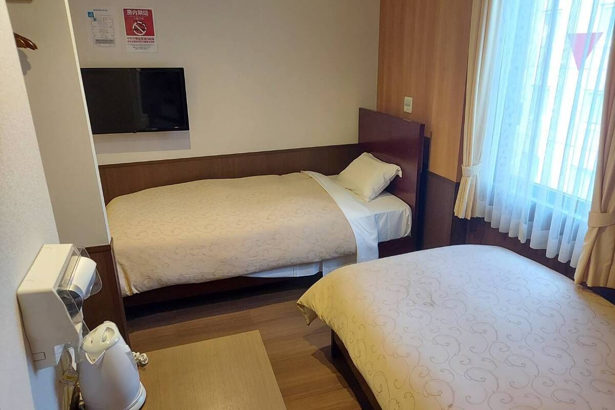 Habitaci ón de hotel con dos camas individuales, televisor montado en la pared, cafetera y ventana con cortinas.
