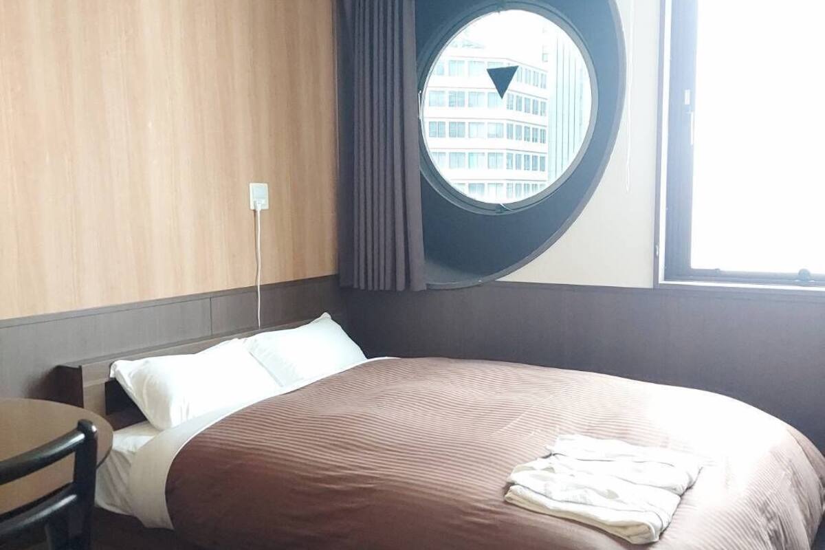 Habitación de hotel con una cama grande, un ventanal redondo con vistas a edificios, un escritorio y una silla.