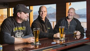Three men sitting at a bar with glasses of beer, wooden walls, and a view of mountains in the background. - Mat-Su Resort (Wasilla)