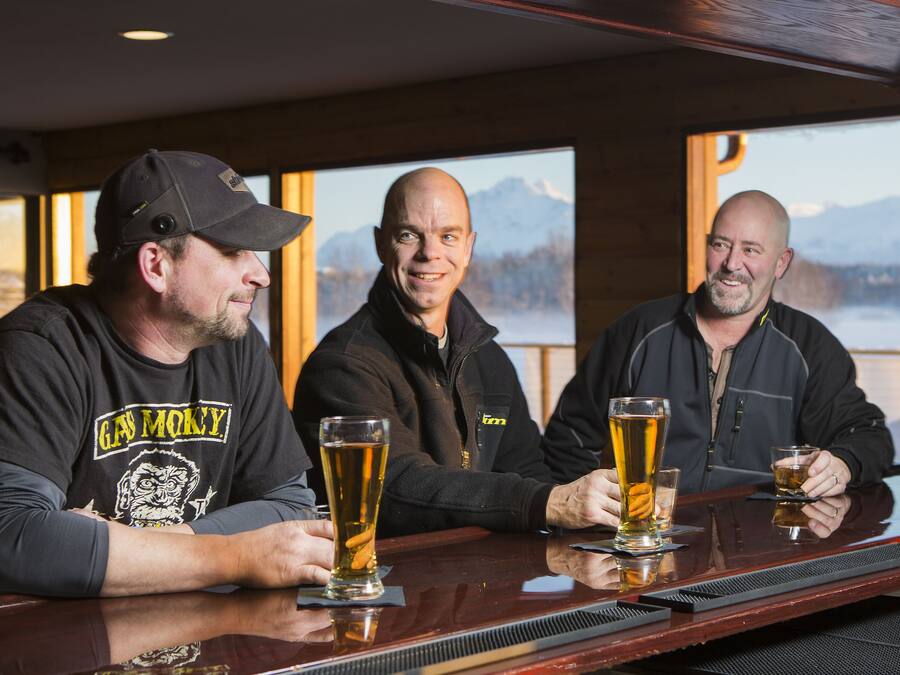 Trois hommes sont assis au bar, devant des verres de bière. Les murs sont en bois et, au fond, on aperçoit des montagnes.