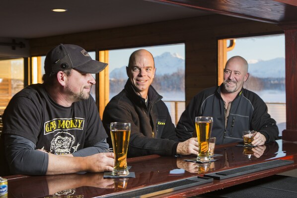 Three men sitting at a bar with glasses of beer, wooden walls, and a view of mountains in the background.